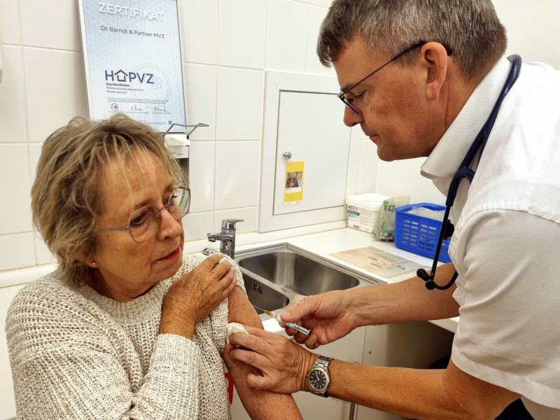 Hausarzt Dr. Matthias Berndt impft Patientin Jutta Blum gegen Grippe in den Oberarm.